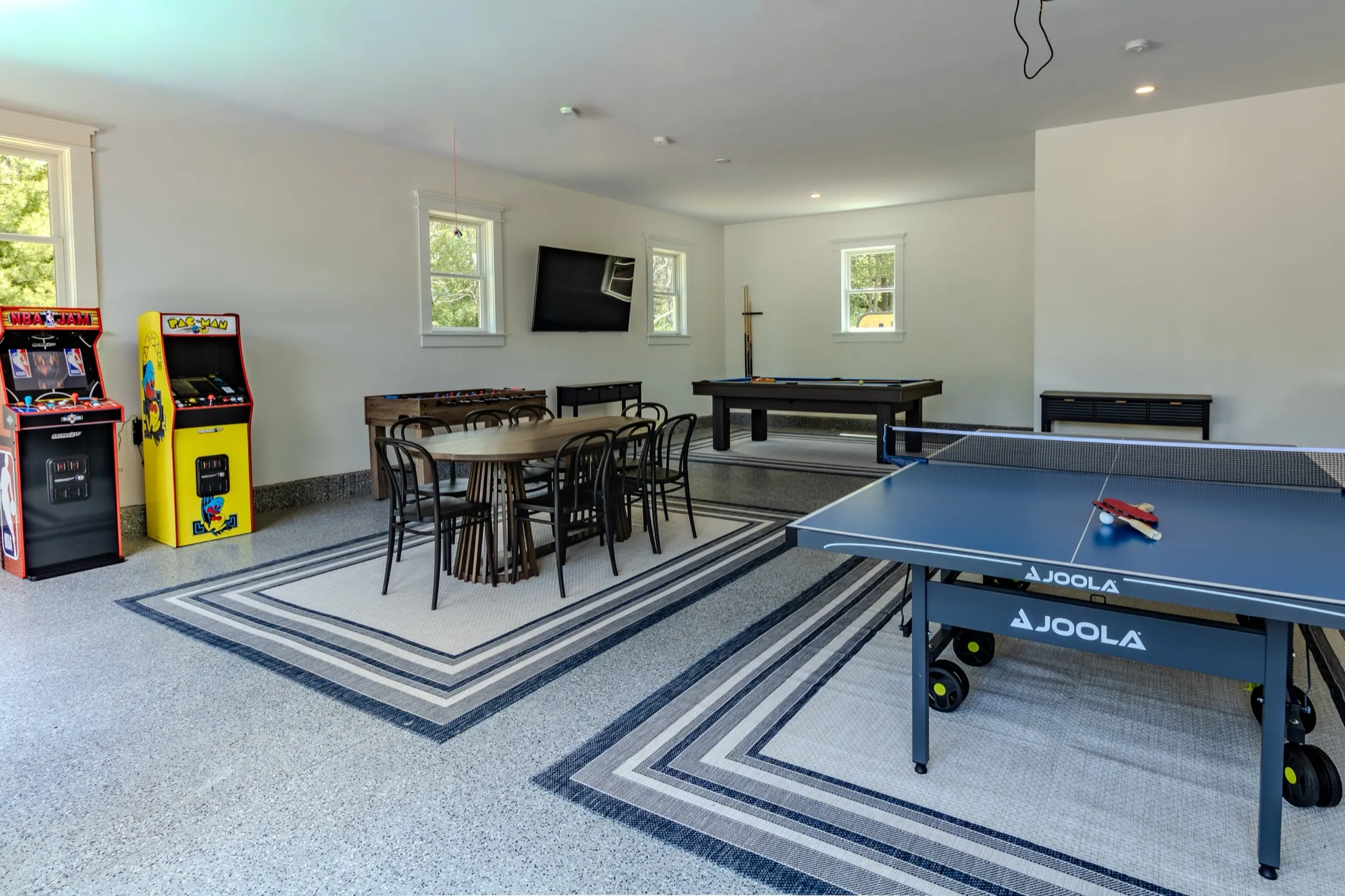Lower-level game room with a ping-pong table, pool table, arcade cabinets, and a dining table on a patterned rug.