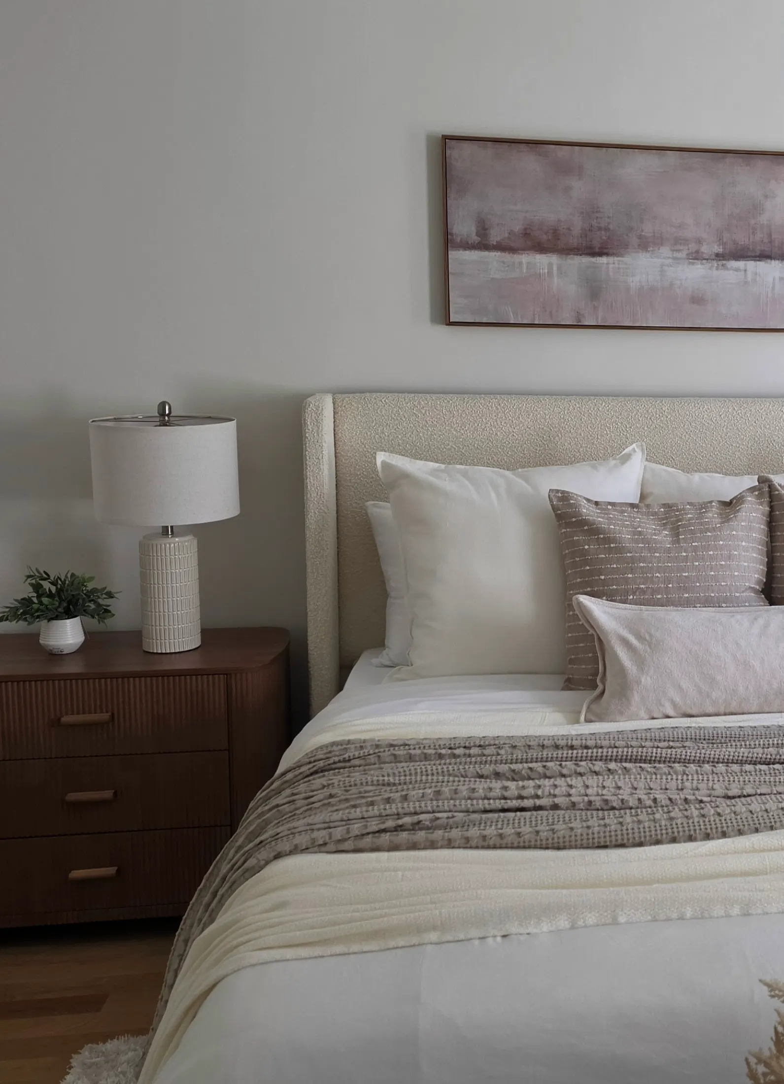 The primary bedroom at 27 Walden Street: a cream boucle headboard, layered white and taupe linens, a woven throw, a fluted walnut nightstand with a textured ceramic lamp, and a soft abstract landscape above the bed.